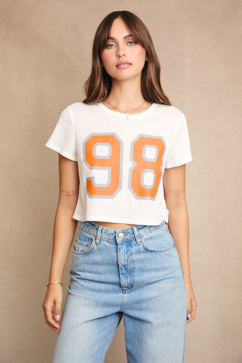 Woman wearing a white t-shirt with orange numbers and blue jeans on a beige background