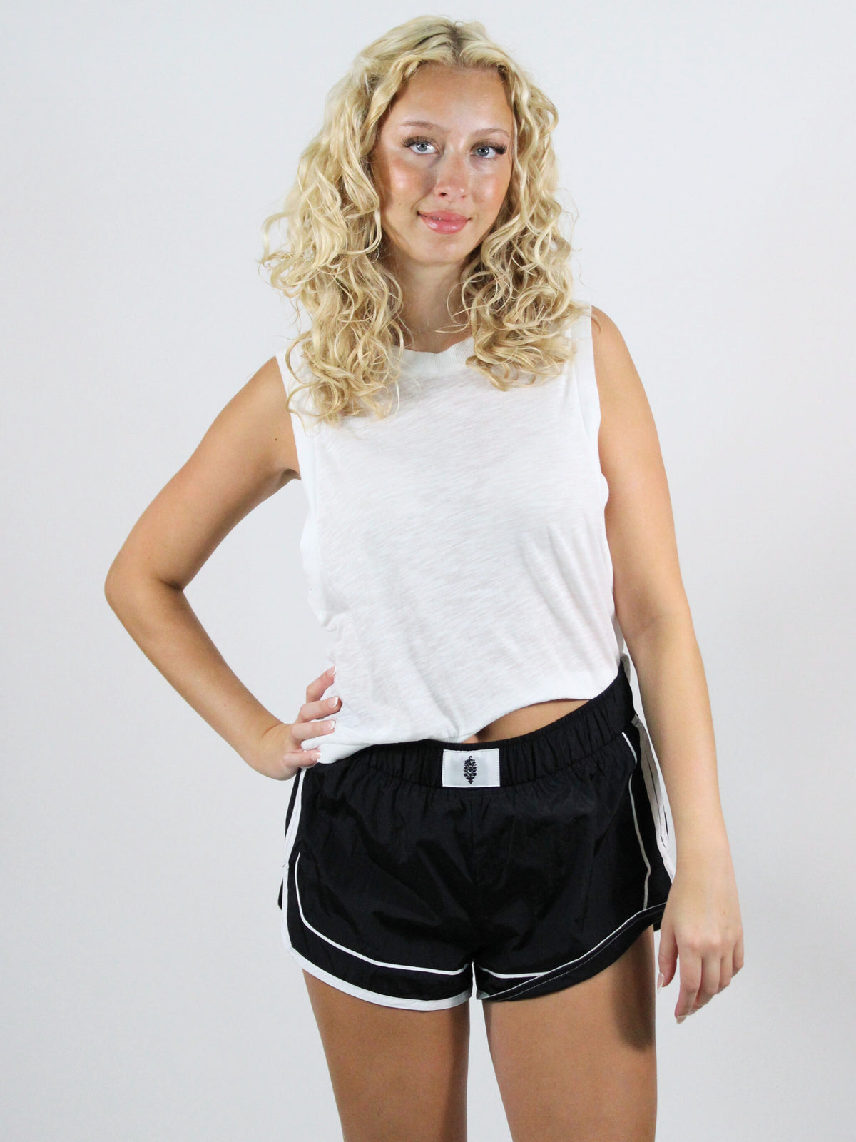Woman wearing a white sleeveless top and black shorts with a logo on a plain background