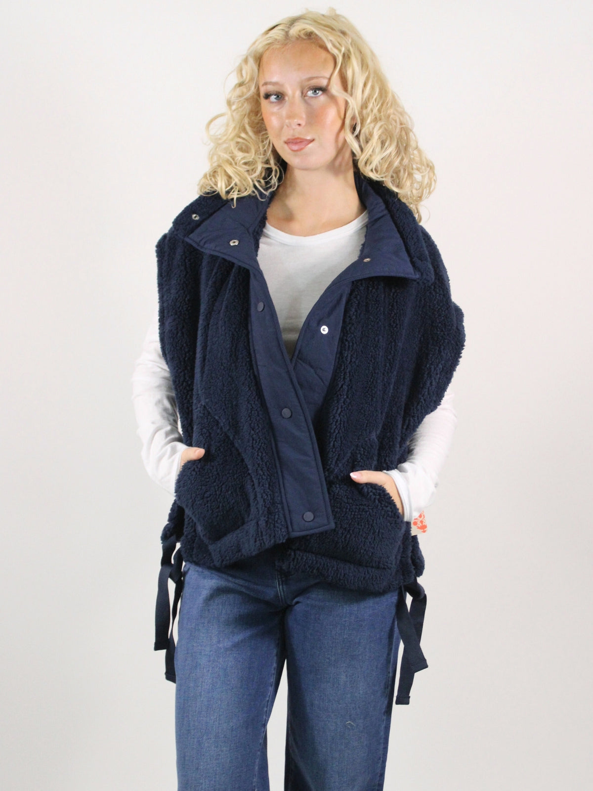 Woman wearing a navy blue vest over a white shirt and blue jeans on a plain background