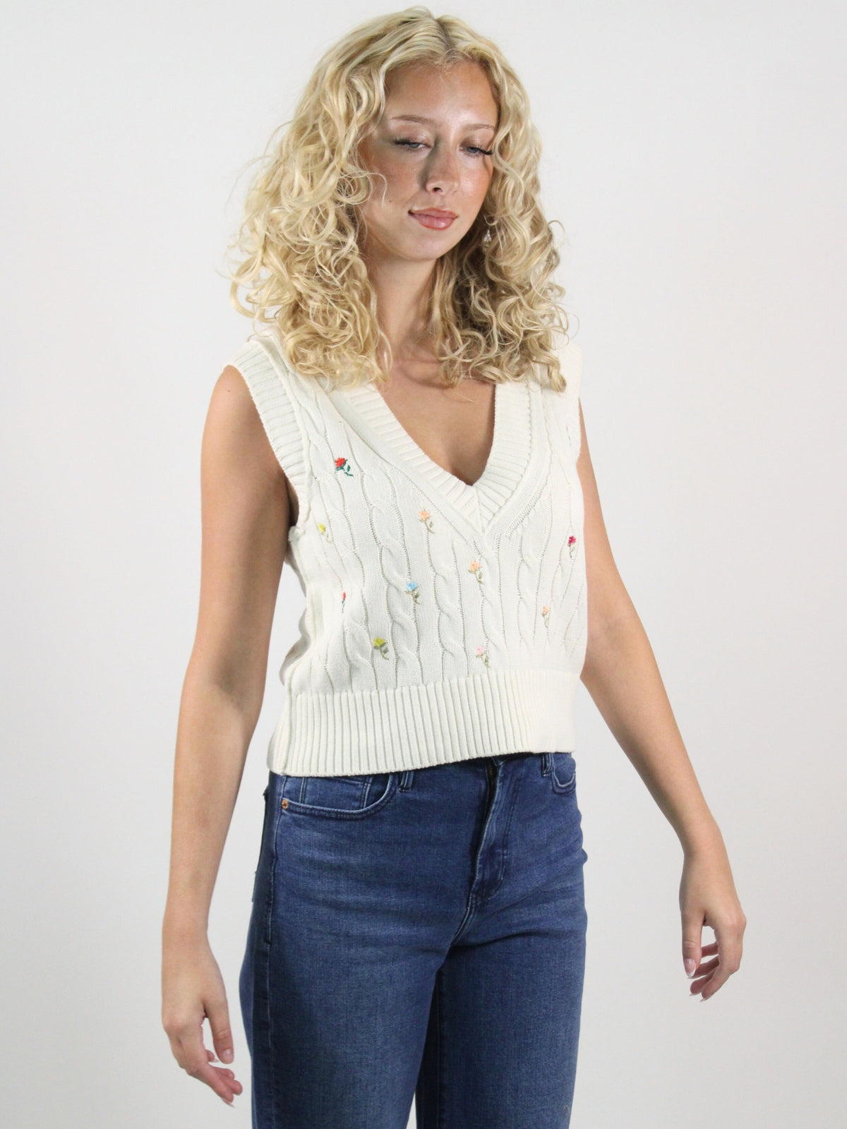 Woman wearing a white sleeveless knitted top with floral patterns on a plain background