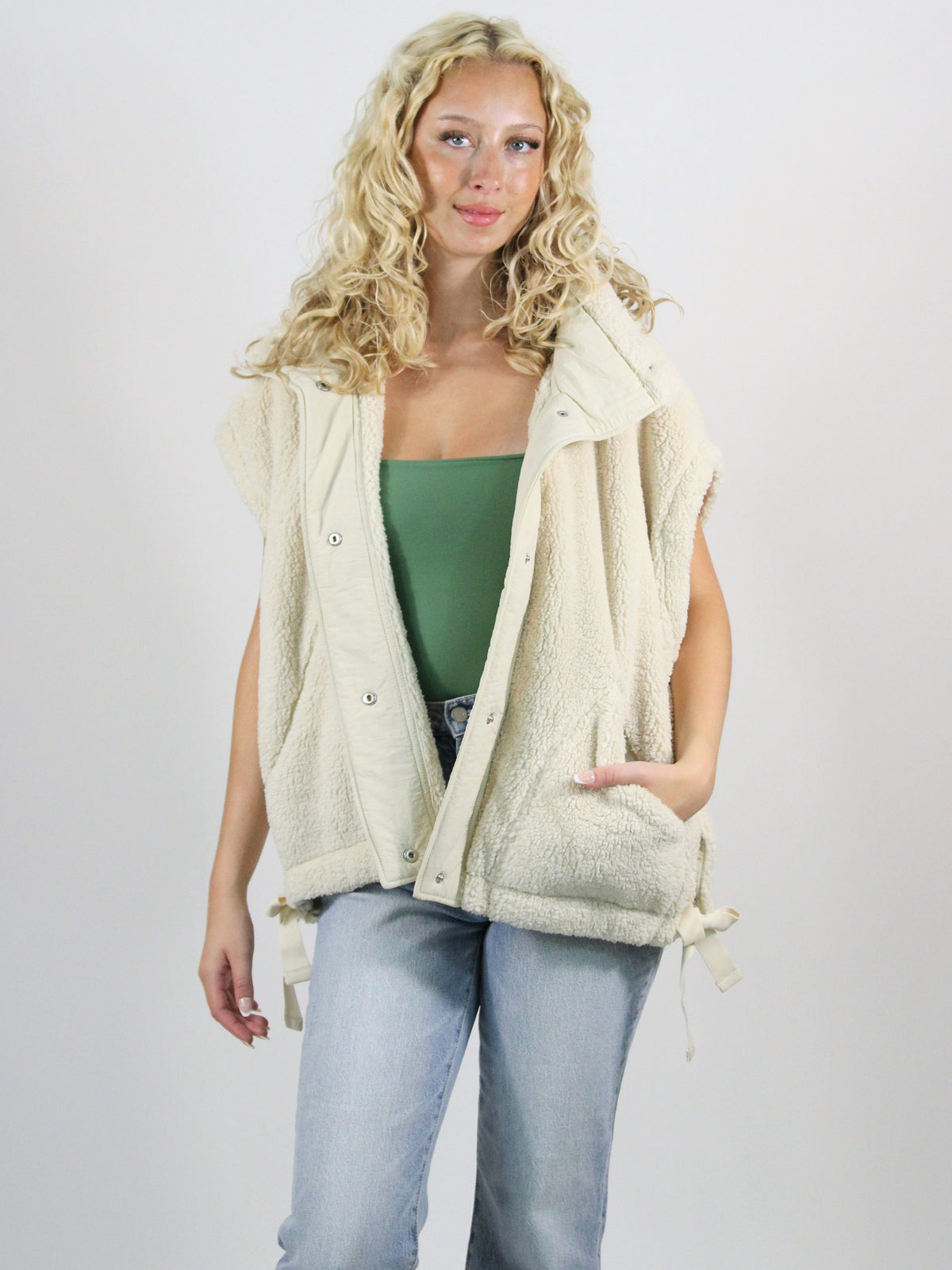 Woman wearing a cream-colored vest over a green top and light blue jeans on a plain background