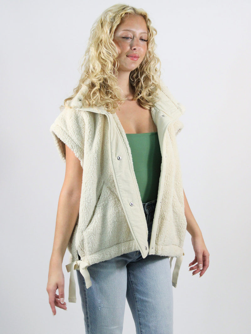 Woman wearing a cream-colored vest over a green top and blue jeans on a white background