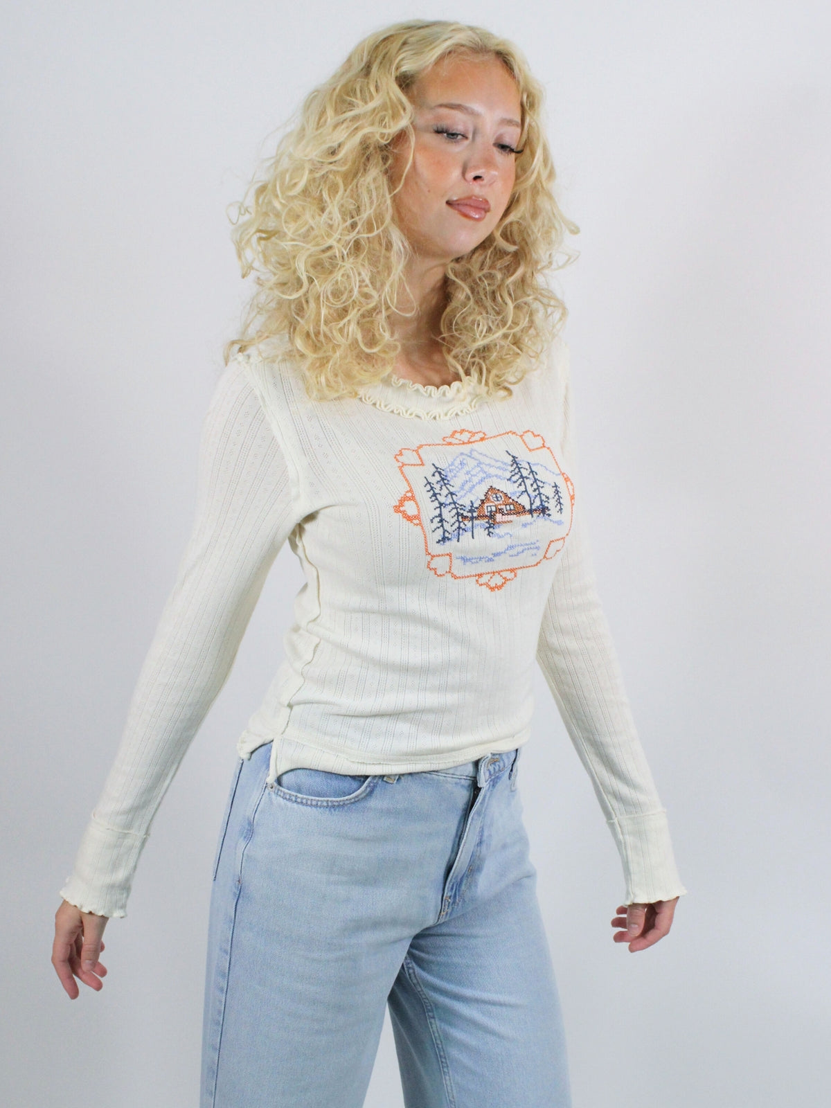 Woman wearing a cream long-sleeve top with a graphic design and light blue jeans on a white background