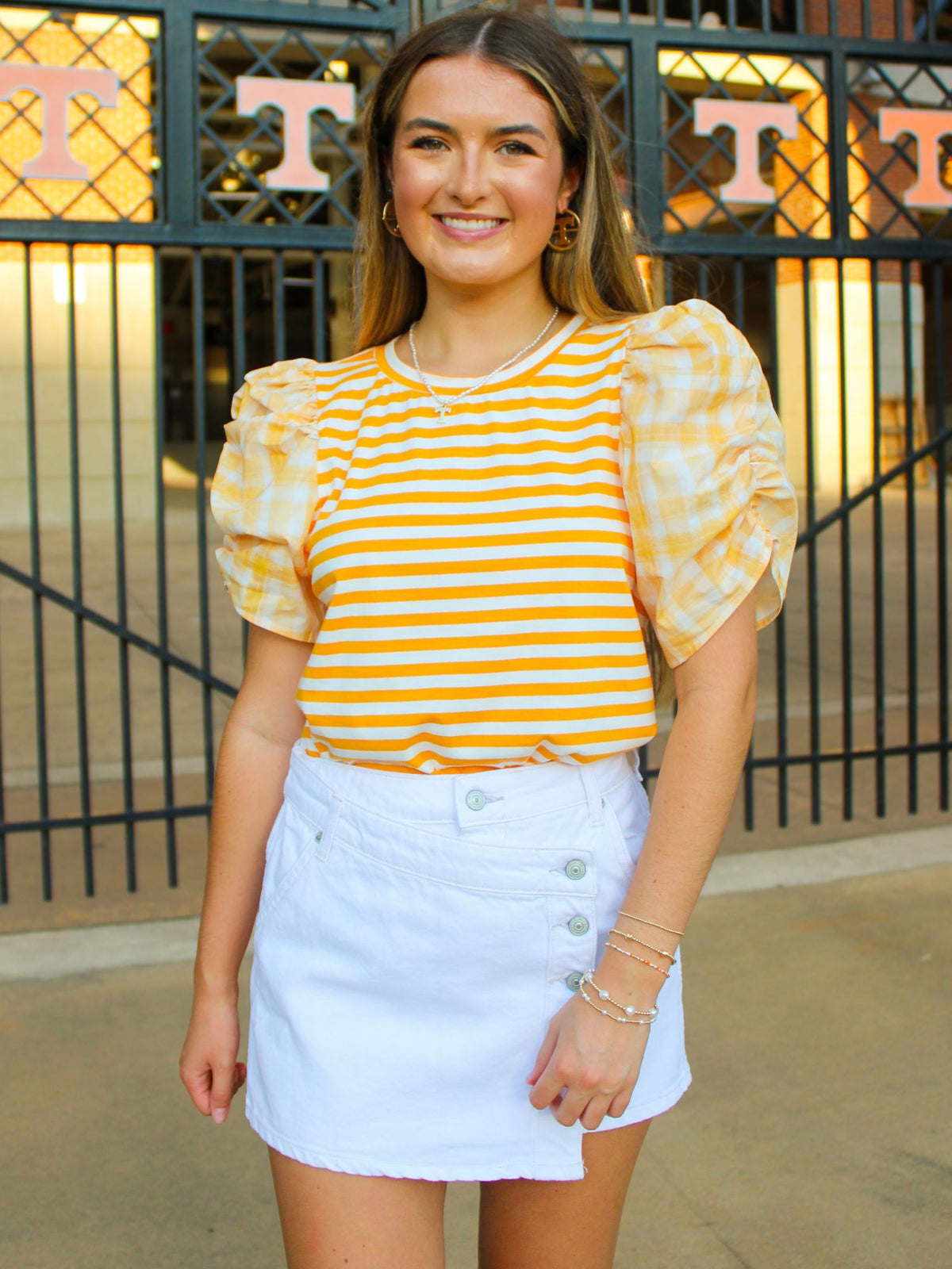 Woman wearing a yellow and white striped shirt with puffed sleeves and a light blue skirt, standing in front of a gate with 'UT' on it.