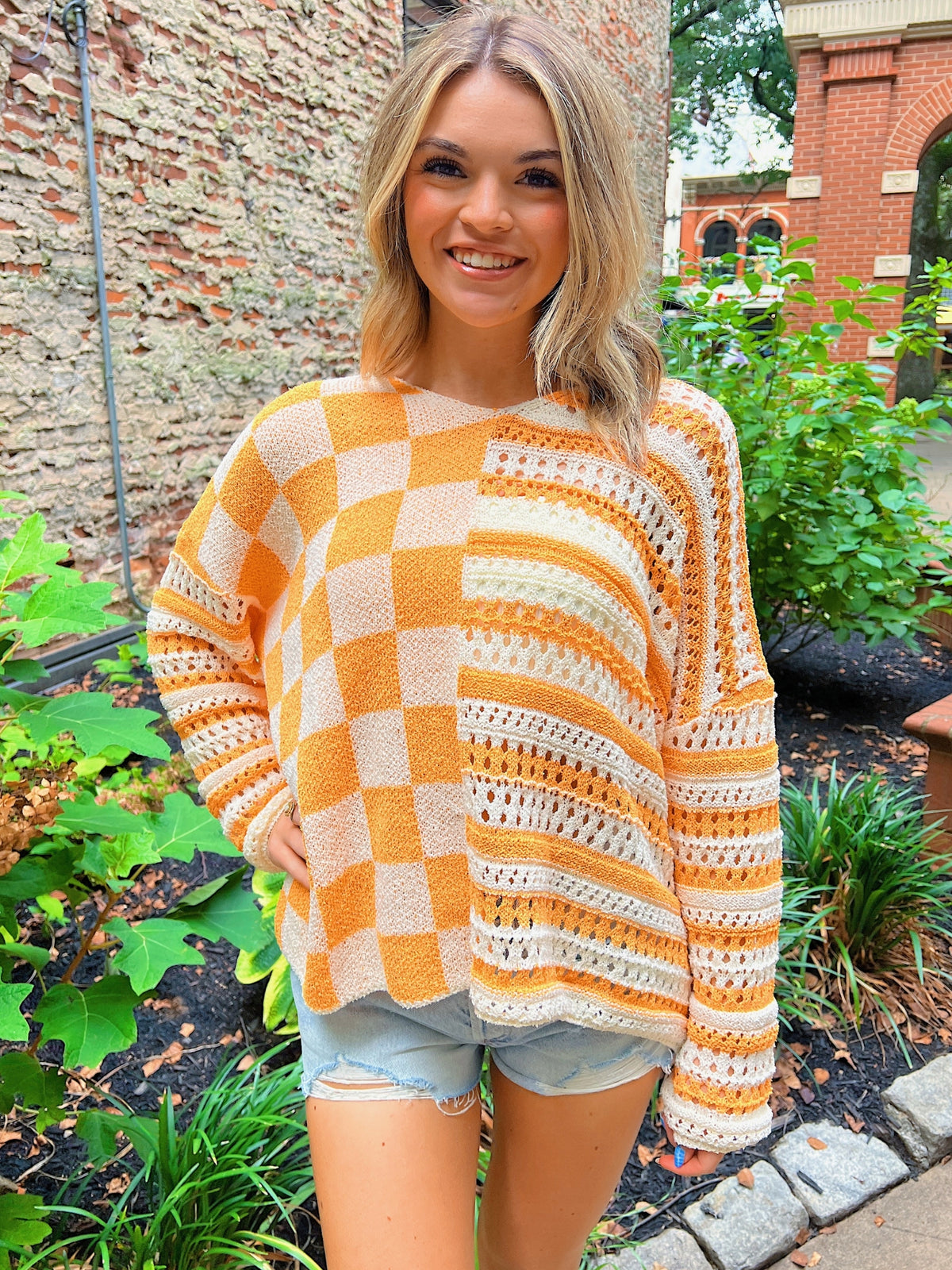 Woman wearing a checkered sweater in an outdoor setting with plants and a brick wall.